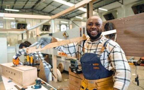 happy-carpenter-wood-enjoy-working-large-wood-workshop-smiling-black-joiner-carrying-pile-wood-making-furniture